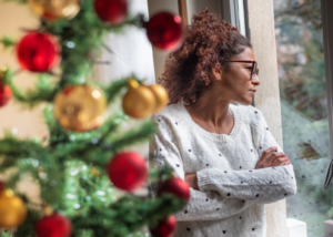 Woman standing next to a Christmas tree, looking out of a window.