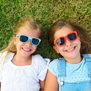Two little girls wearing sunglasses laying on the grass, looking up at the sky.