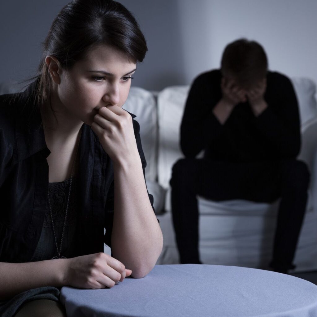 Woman sitting at table with man in the background sitting on couch.